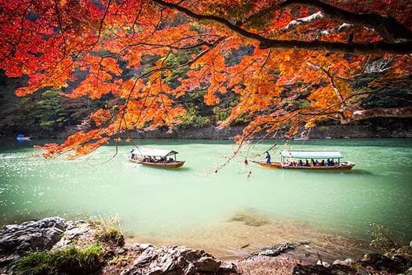 Boats on a jade-green river in Kyoto framed by red autumn maple leaves