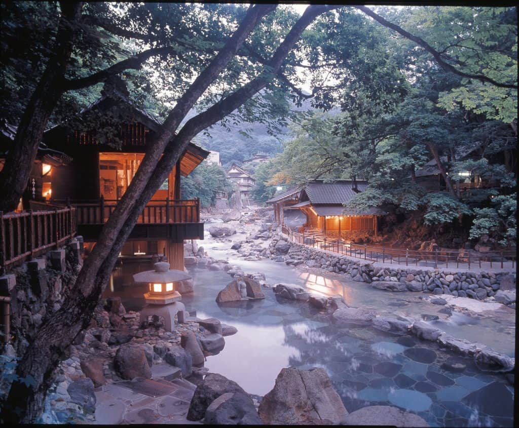 Outdoor stone hot spring pools beside a river in a wooded valley with wooden inns lit at dusk