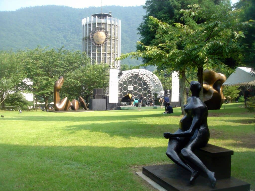 A green lawn with modern sculptures and a tall glass tower at an open-air art park in Hakone