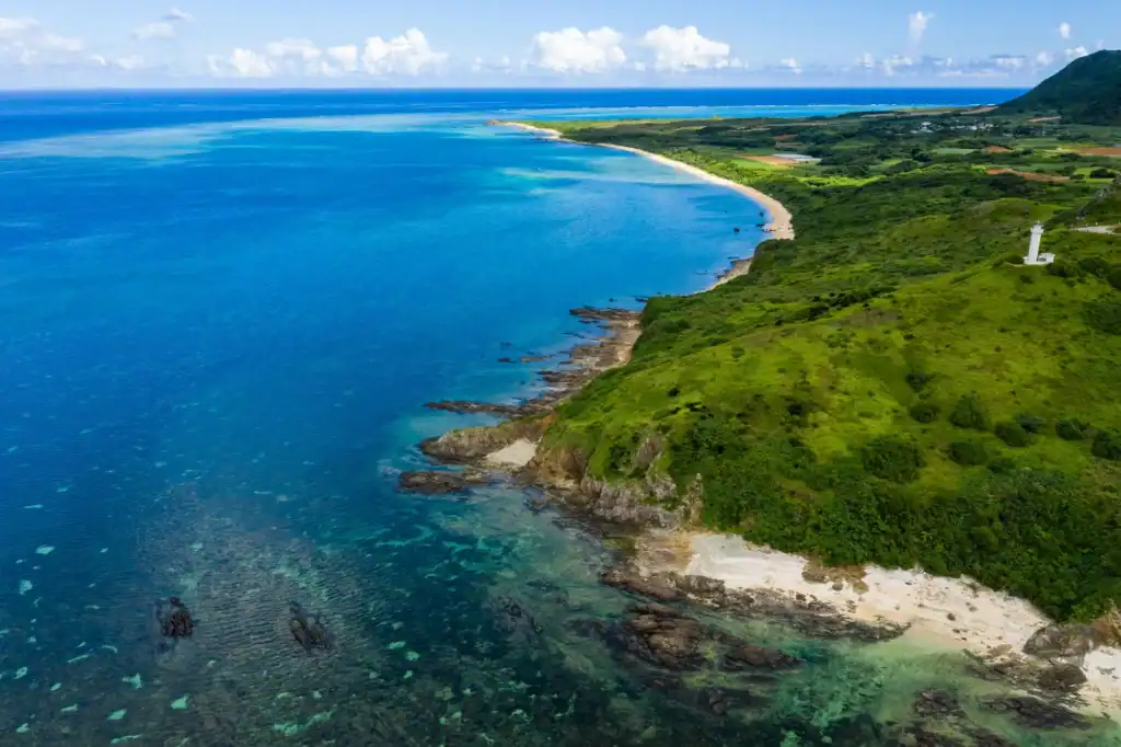 An aerial view of an Okinawan coastline with turquoise shallows, white sand and green headlands