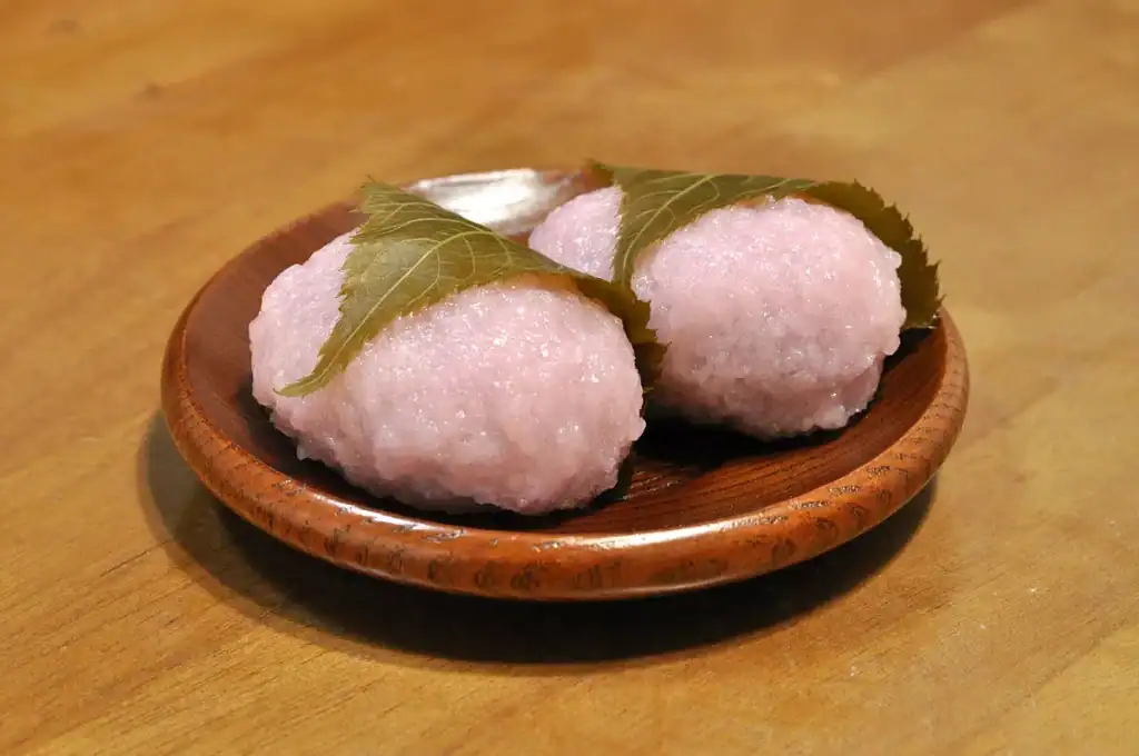 Two pink sakura mochi rice cakes wrapped in green cherry leaves on a wooden plate