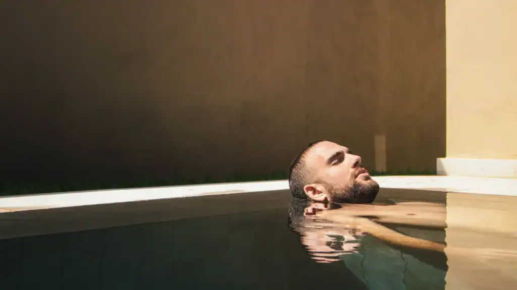 A young man resting his head on the rim of a dimly lit indoor bath, eyes closed