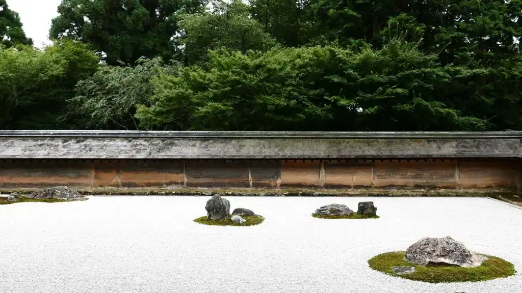 A dry garden of raked white gravel with low moss islands and a few stones before an earthen wall