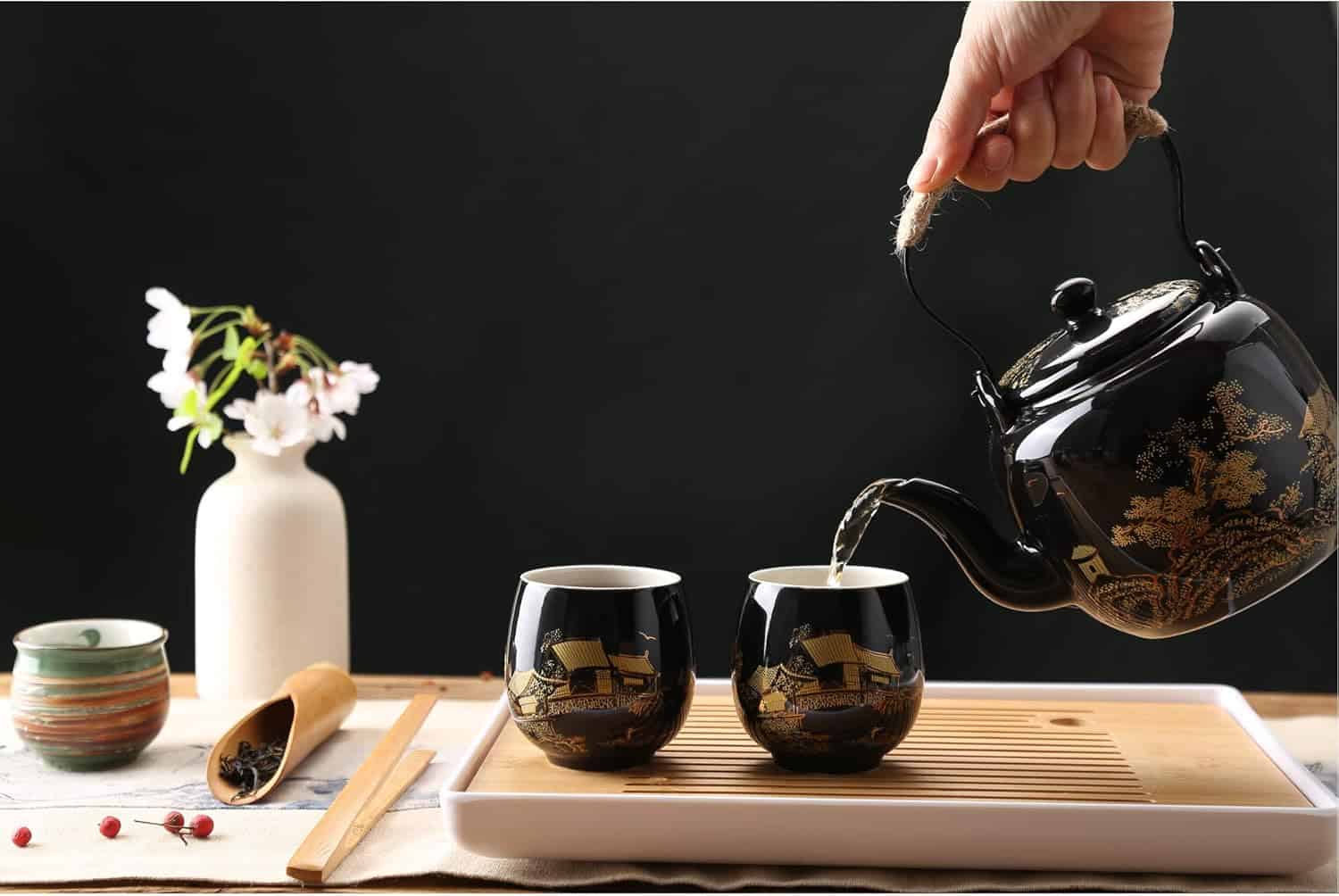 A hand pouring tea from a black lacquered teapot into two cups on a wooden tray
