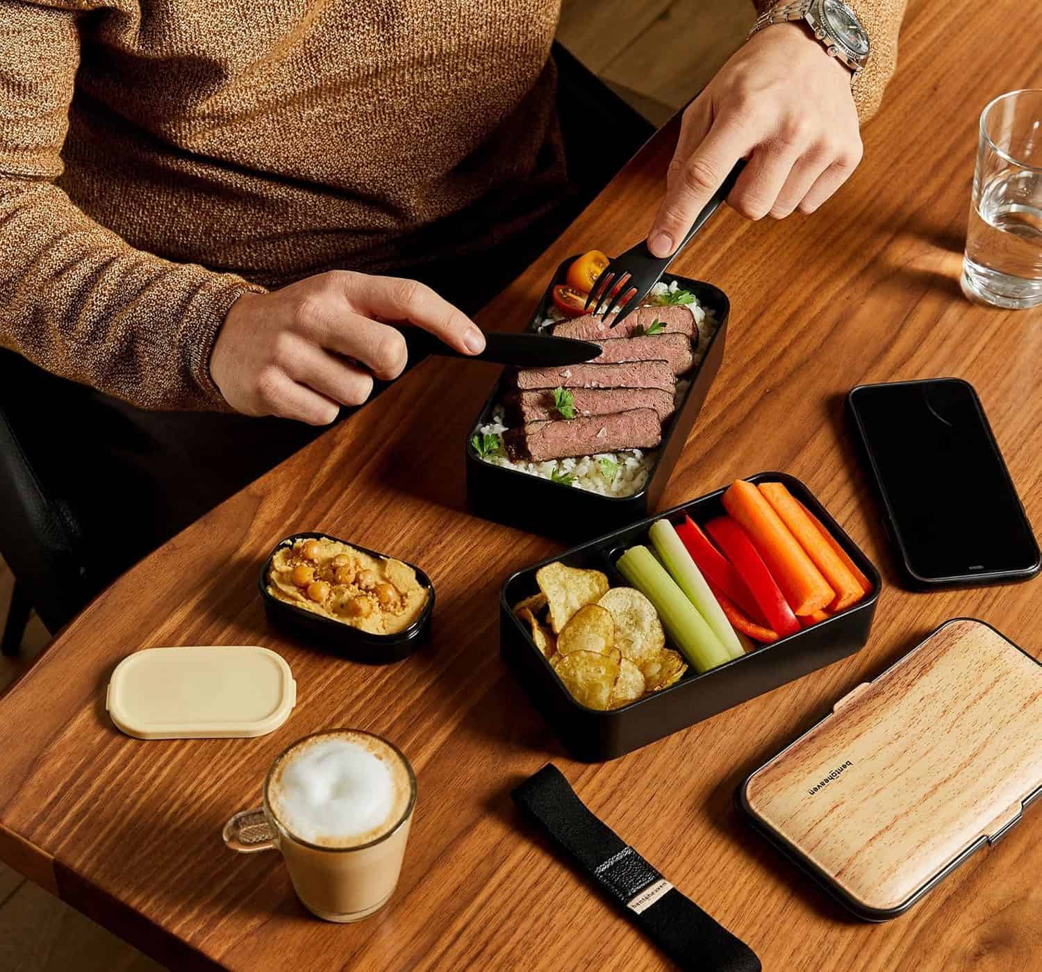 A bento box of beef, vegetables and snacks beside a latte and a phone on a wooden table