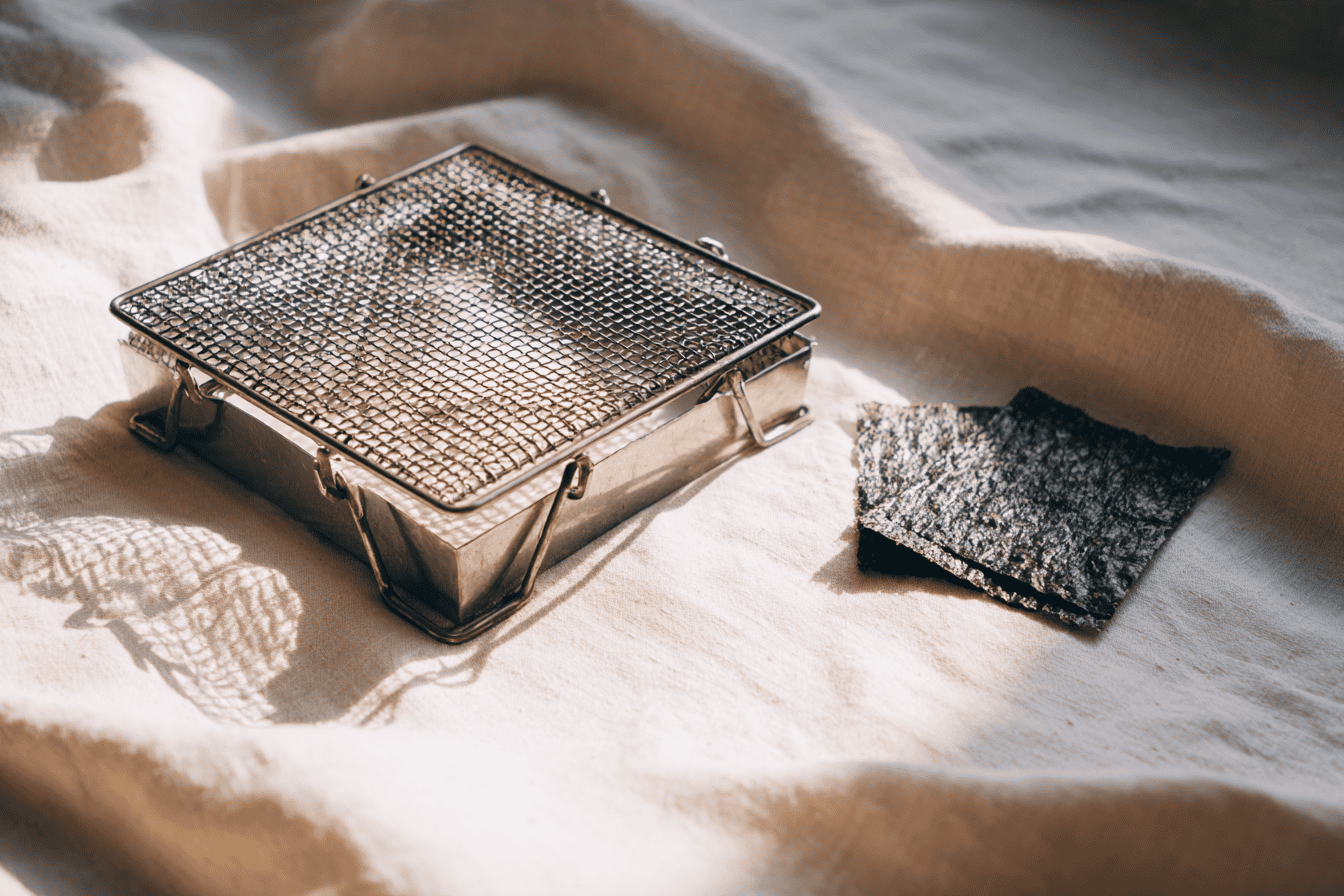 A square Japanese yakimi-ami wire grilling net on cream linen, beside a crisp sheet of toasted nori
