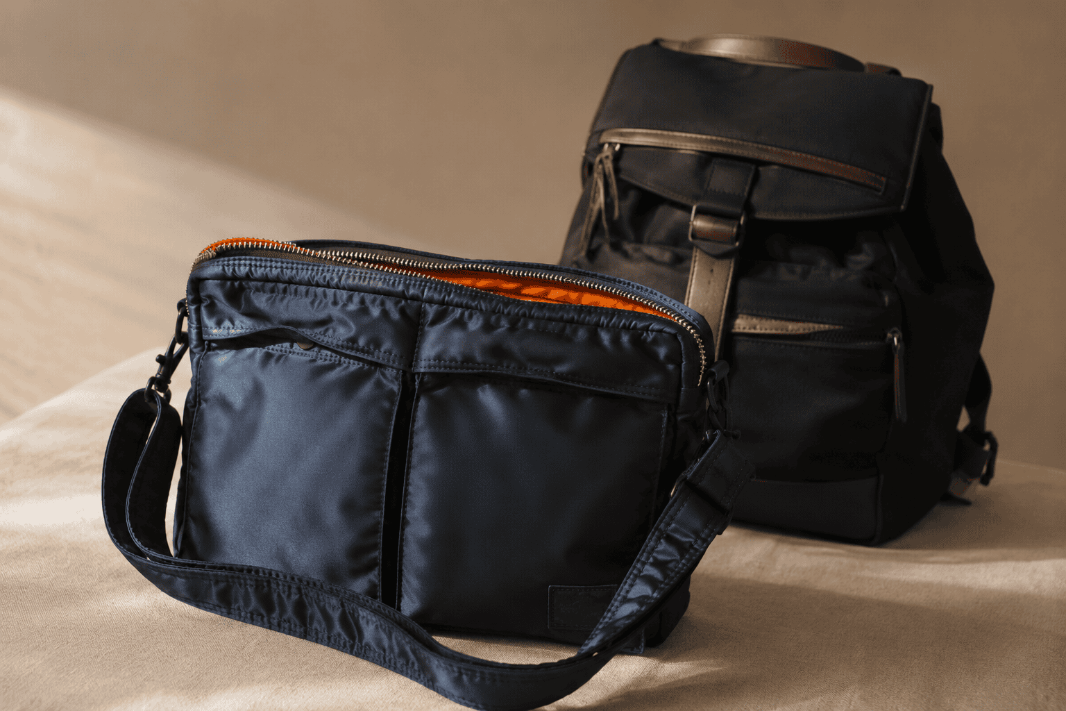 A Porter-Yoshida Tanker bag in iron blue beside a black Master-piece backpack on a cream surface