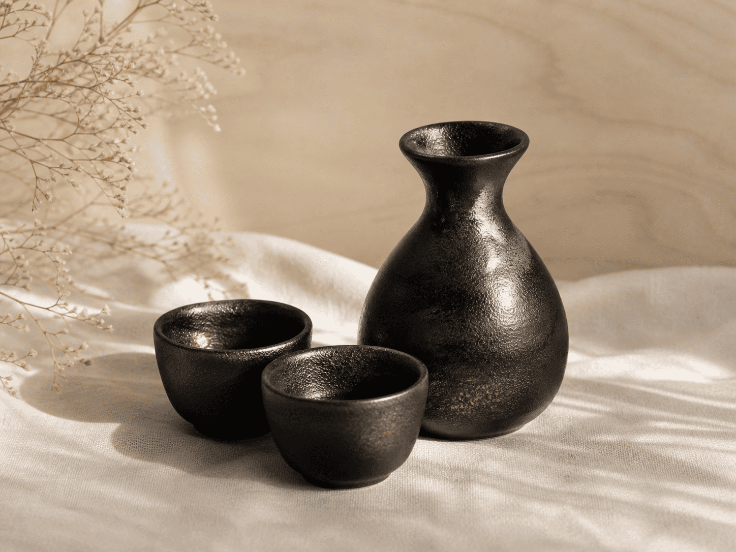 A black ceramic Mino-ware sake set on cream linen — tokkuri carafe and ochoko cups
