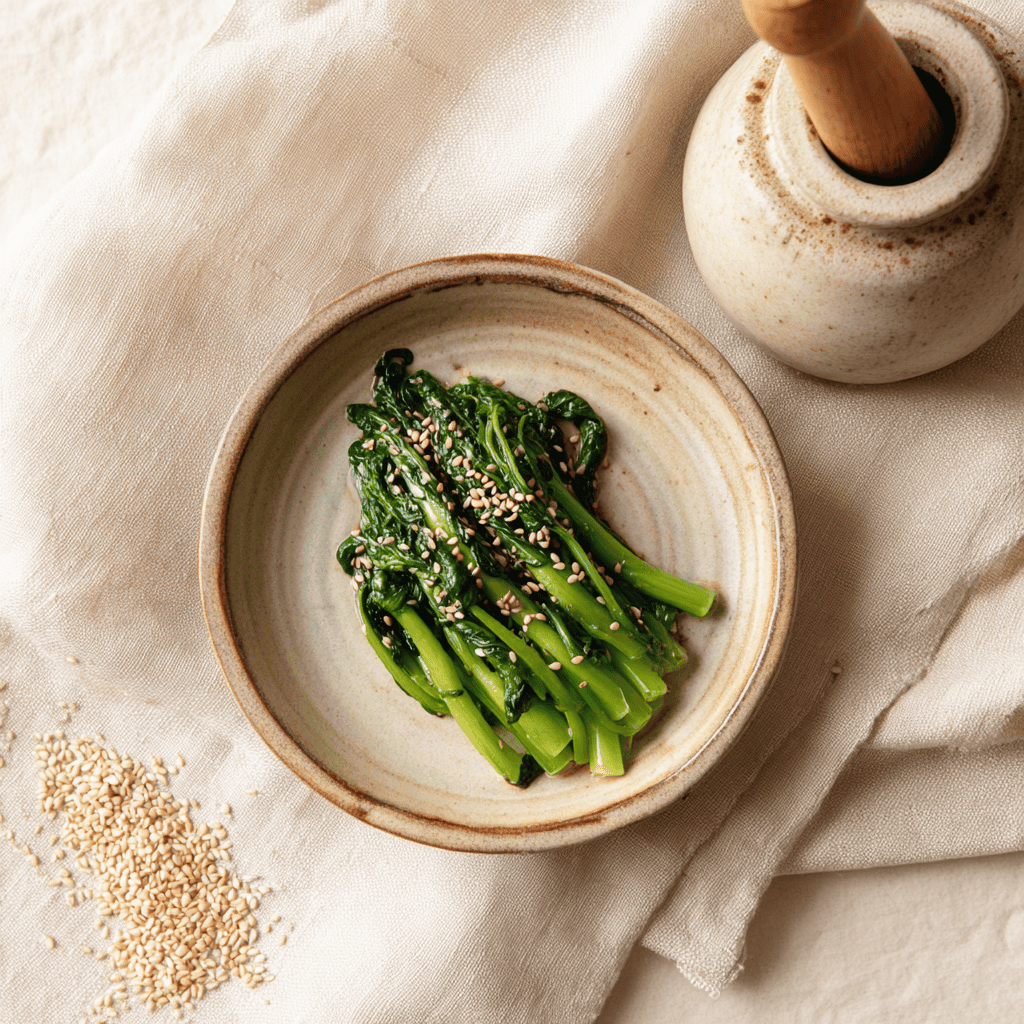 A small ceramic bowl of bright green Japanese spinach gomaae dressed in nutty toasted sesame paste, plated minimally on cream linen