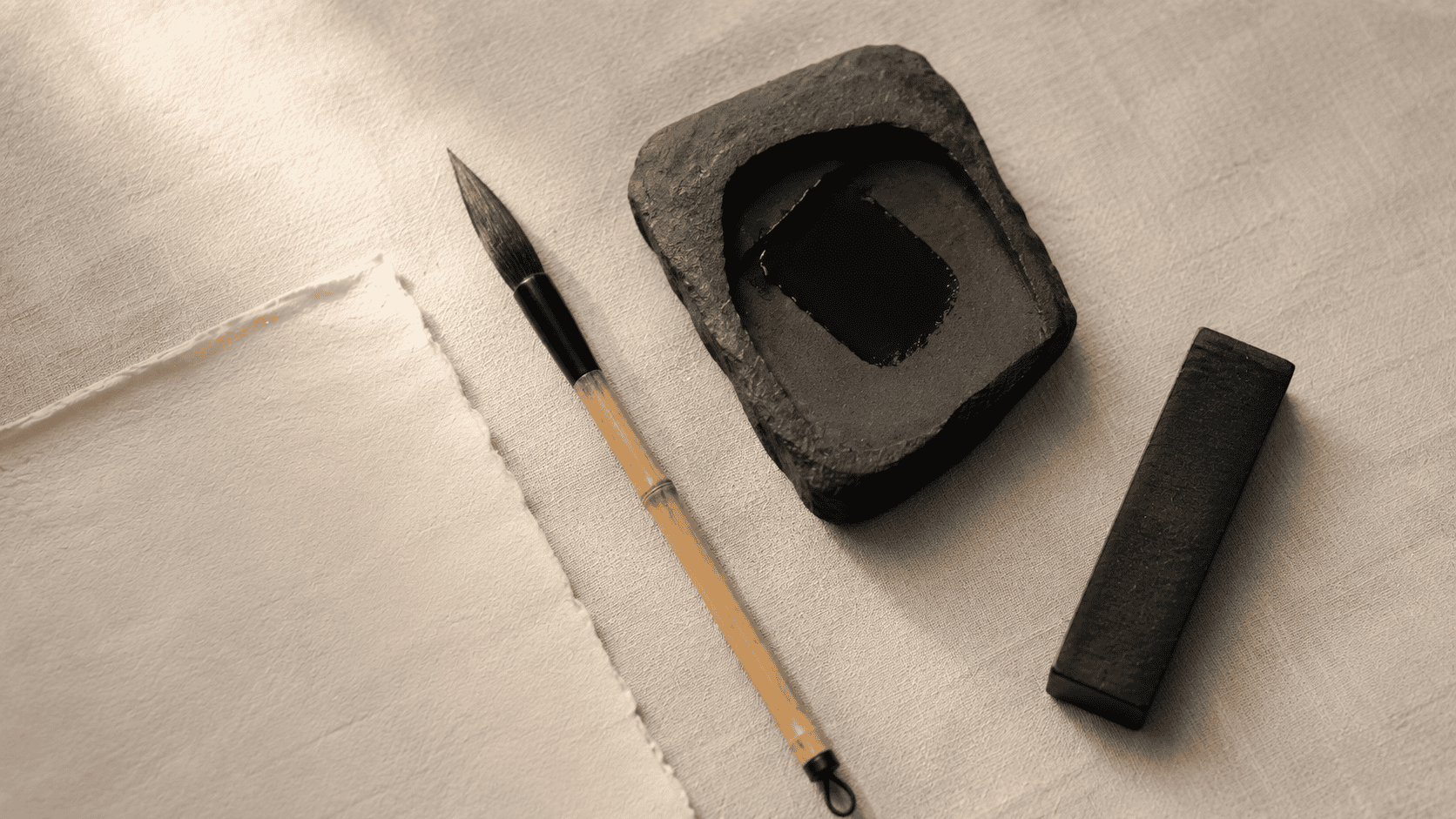 An overhead still life on cream linen of a bamboo-handled calligraphy brush, a black ink stick, a slate inkstone, and a sheet of textured washi paper
