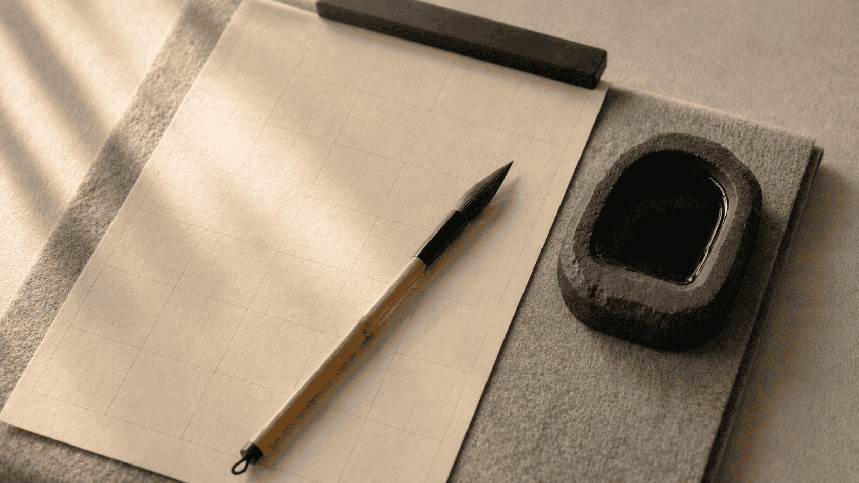A calligraphy brush resting on a sheet of cream practice paper beside a small inkstone, soft daylight from the left, a felt mat beneath
