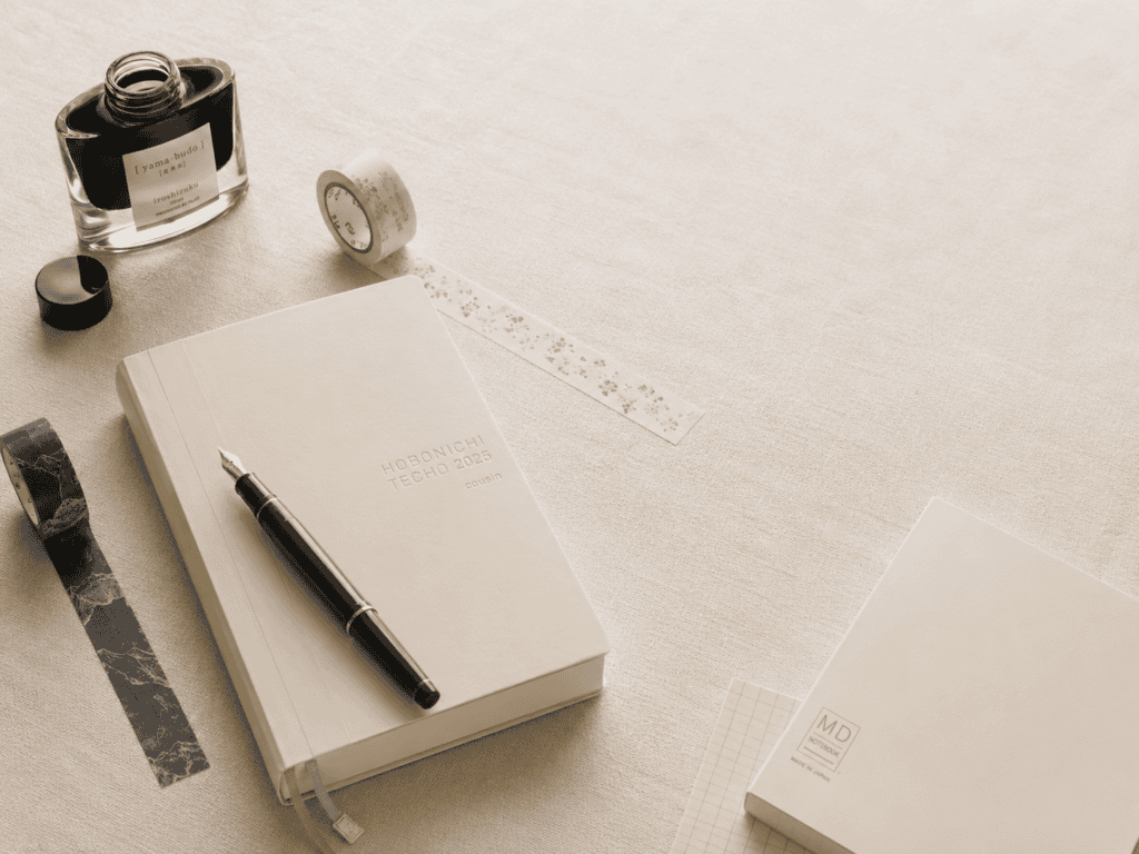A cream notebook with a fountain pen, a bottle of ink and washi tape on a linen desk