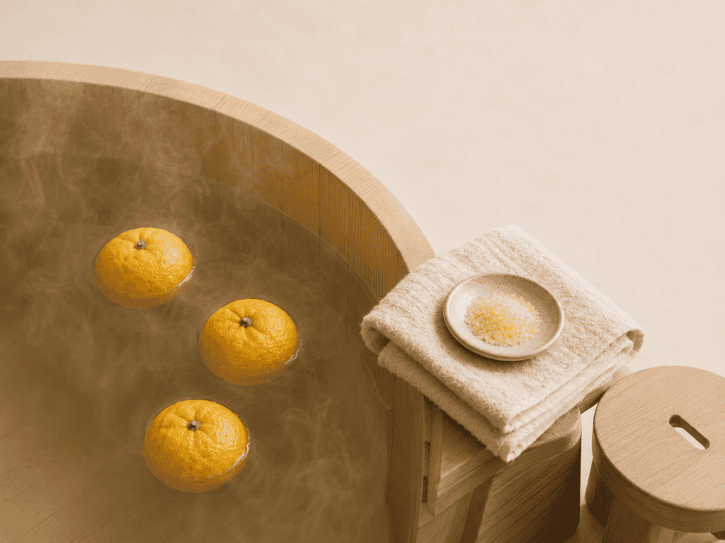 Whole yuzu citrus fruits floating in a round wooden bath beside a folded towel and bath salts