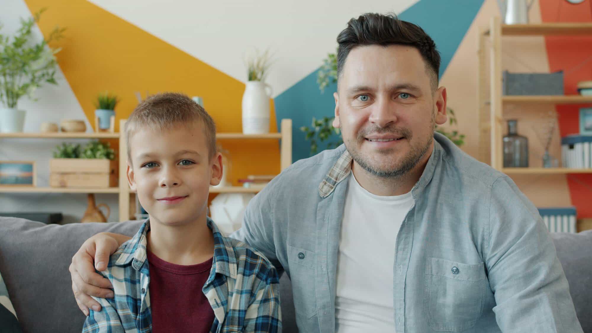 A smiling father with his arm around his young son on a sofa at home