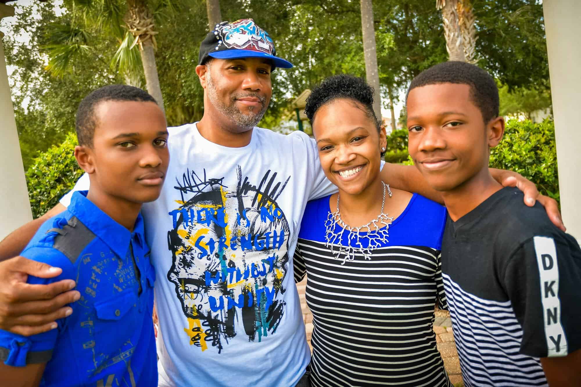 A smiling father with his wife and two teenage sons standing together outdoors on a sunny day