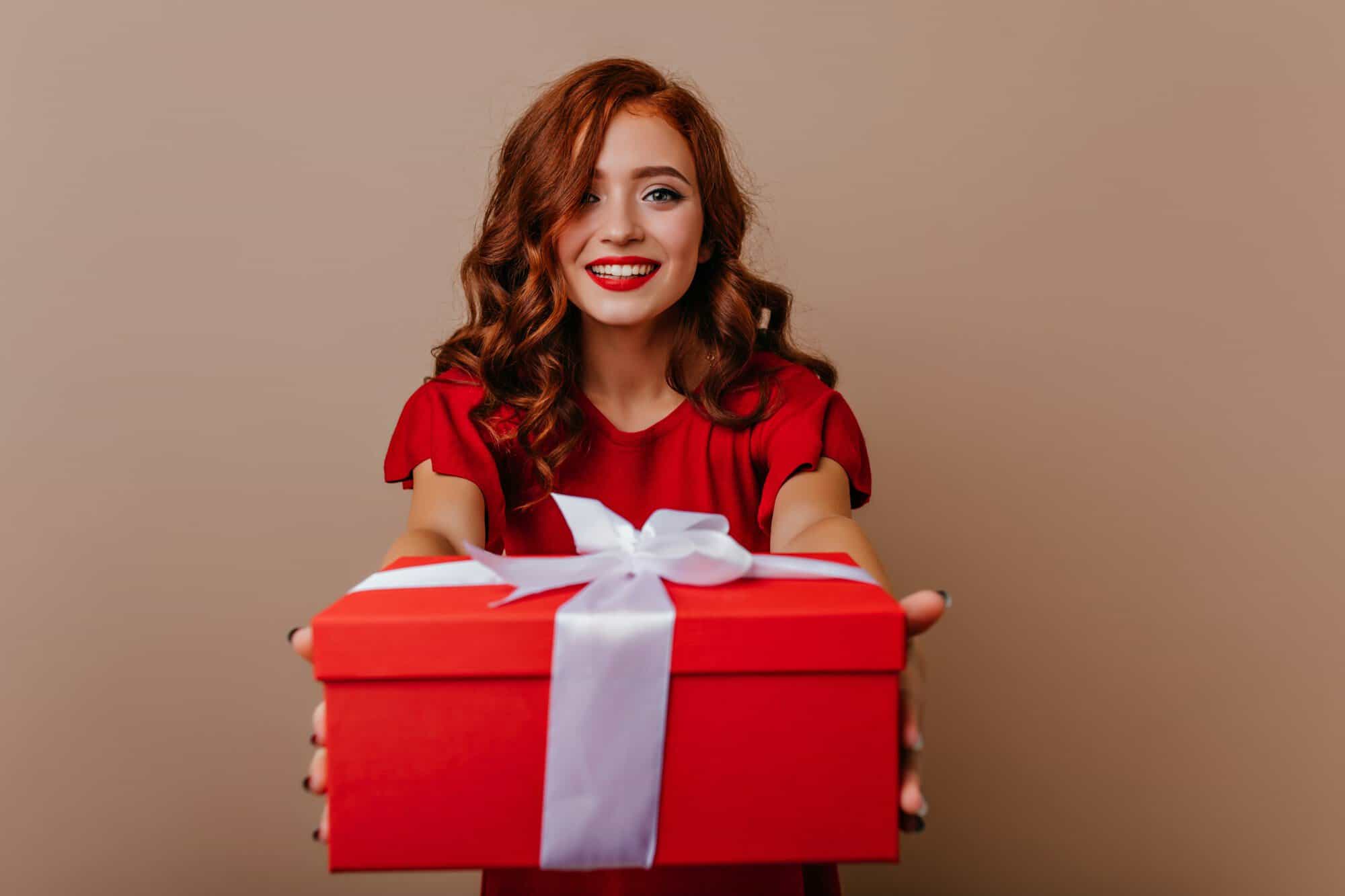 A smiling woman in red holding out a red gift box tied with a white ribbon