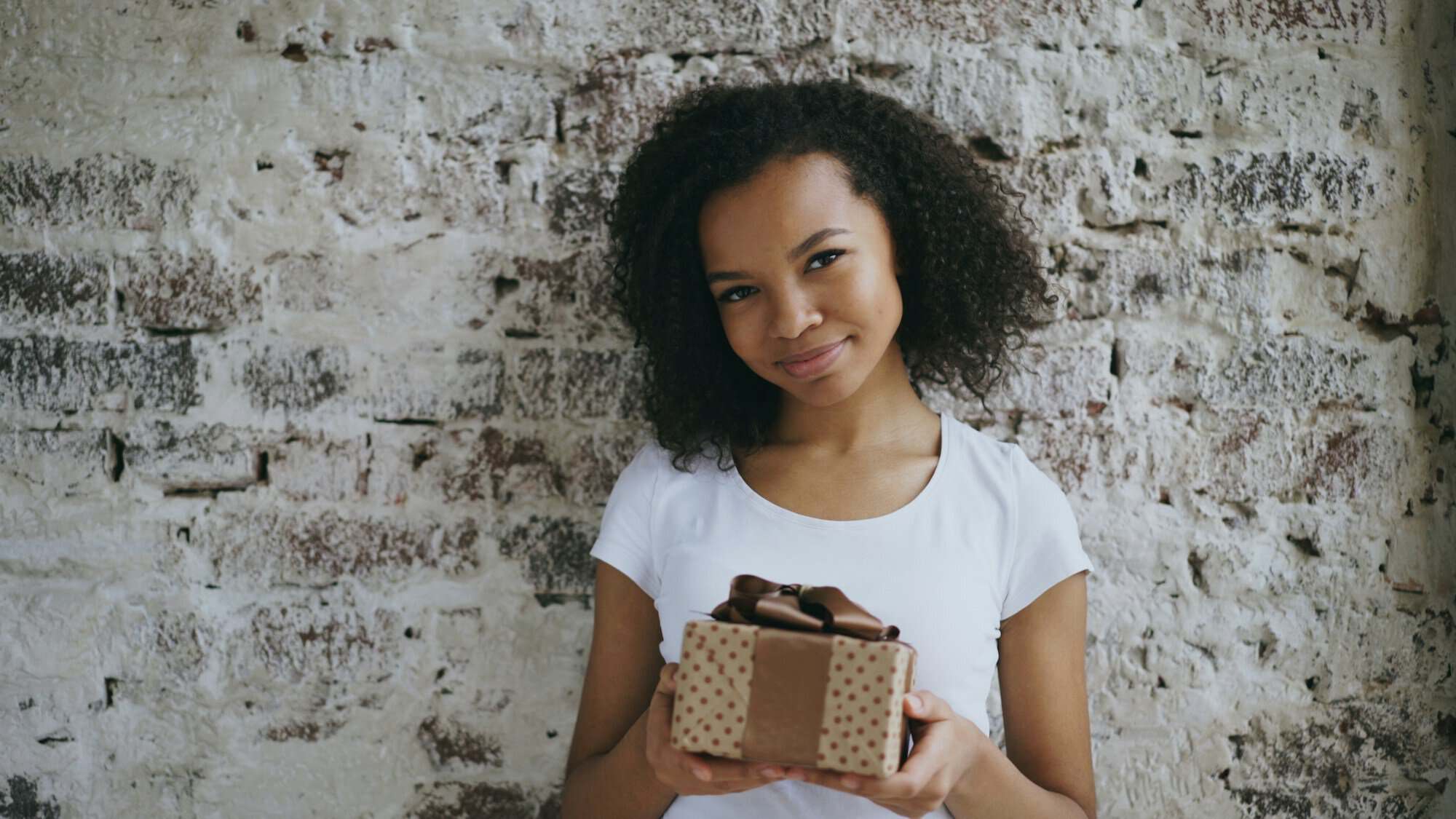 A smiling young woman holding a small wrapped gift box against a whitewashed wall, ready to give one of these Japanese gifts for grandparents