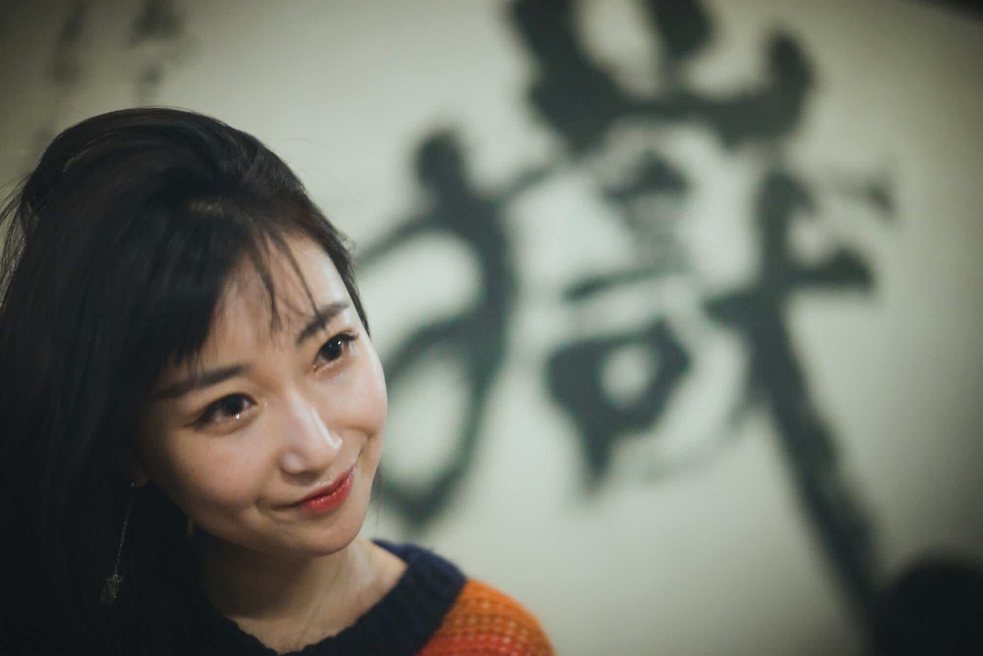 A smiling young woman looking toward the camera in front of a wall painted with large brush characters