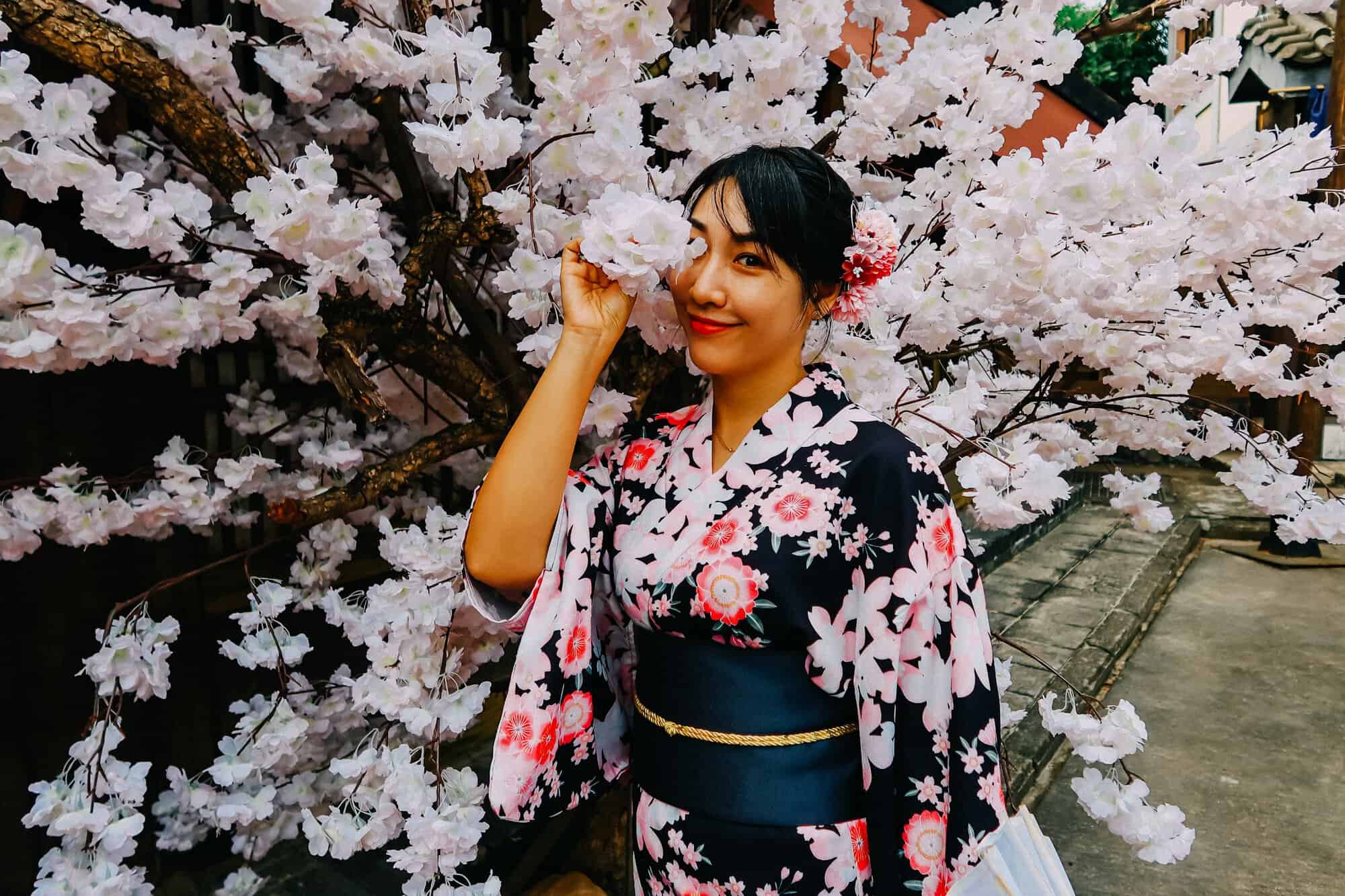 A smiling woman in a floral yukata holding a blossom beside a flowering tree