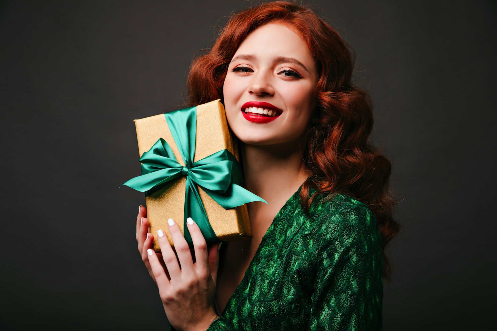 A smiling woman with red curls holding a gift box tied with a green ribbon