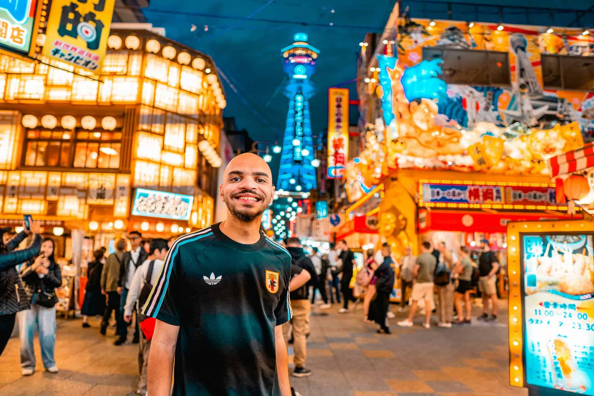 A smiling man standing in a neon-lit Osaka street at night with Tsutenkaku Tower behind him