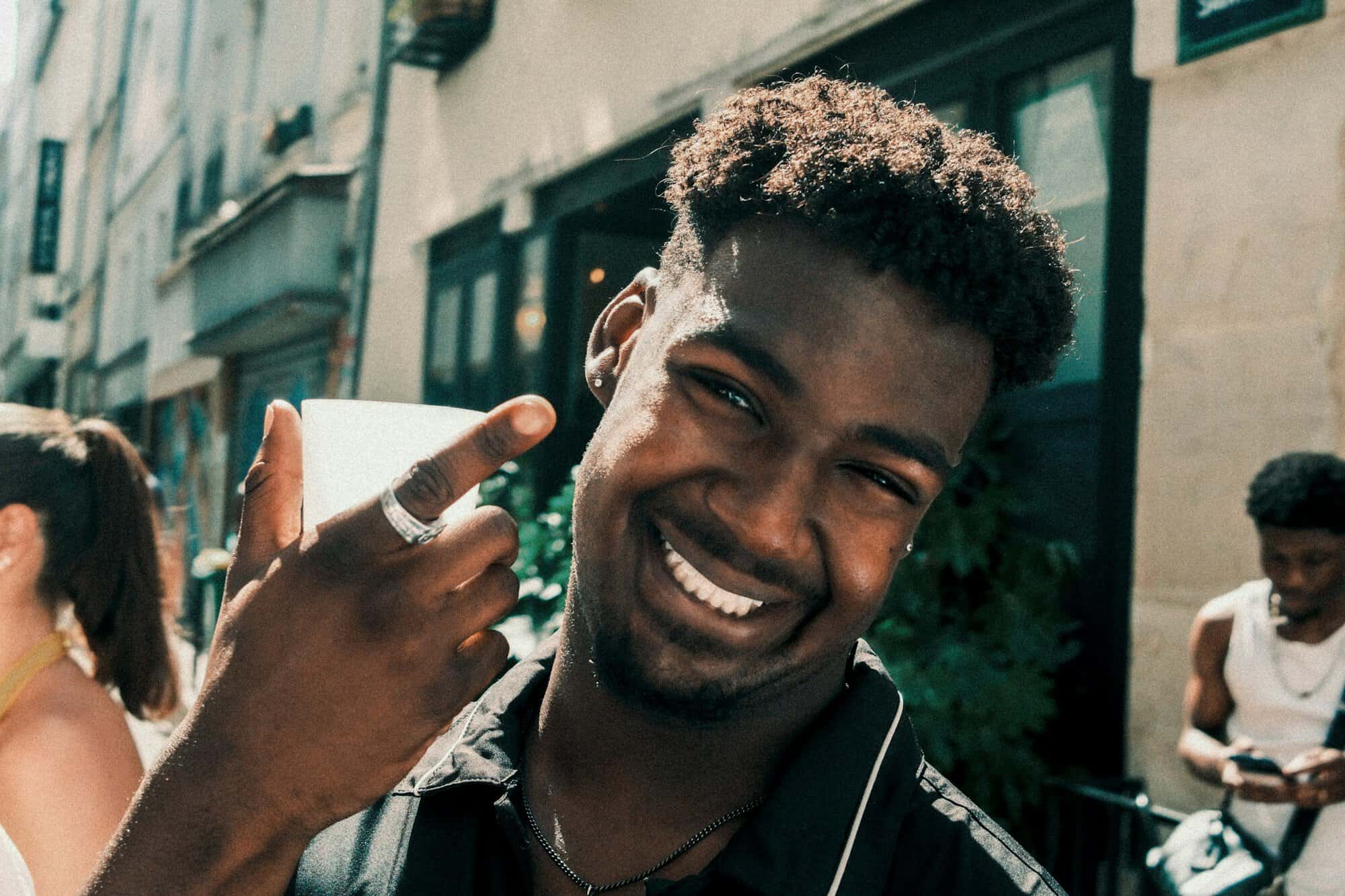 A smiling young man holds up a coffee cup on a sunny street, the kind of partner Japanese gifts for him suit best