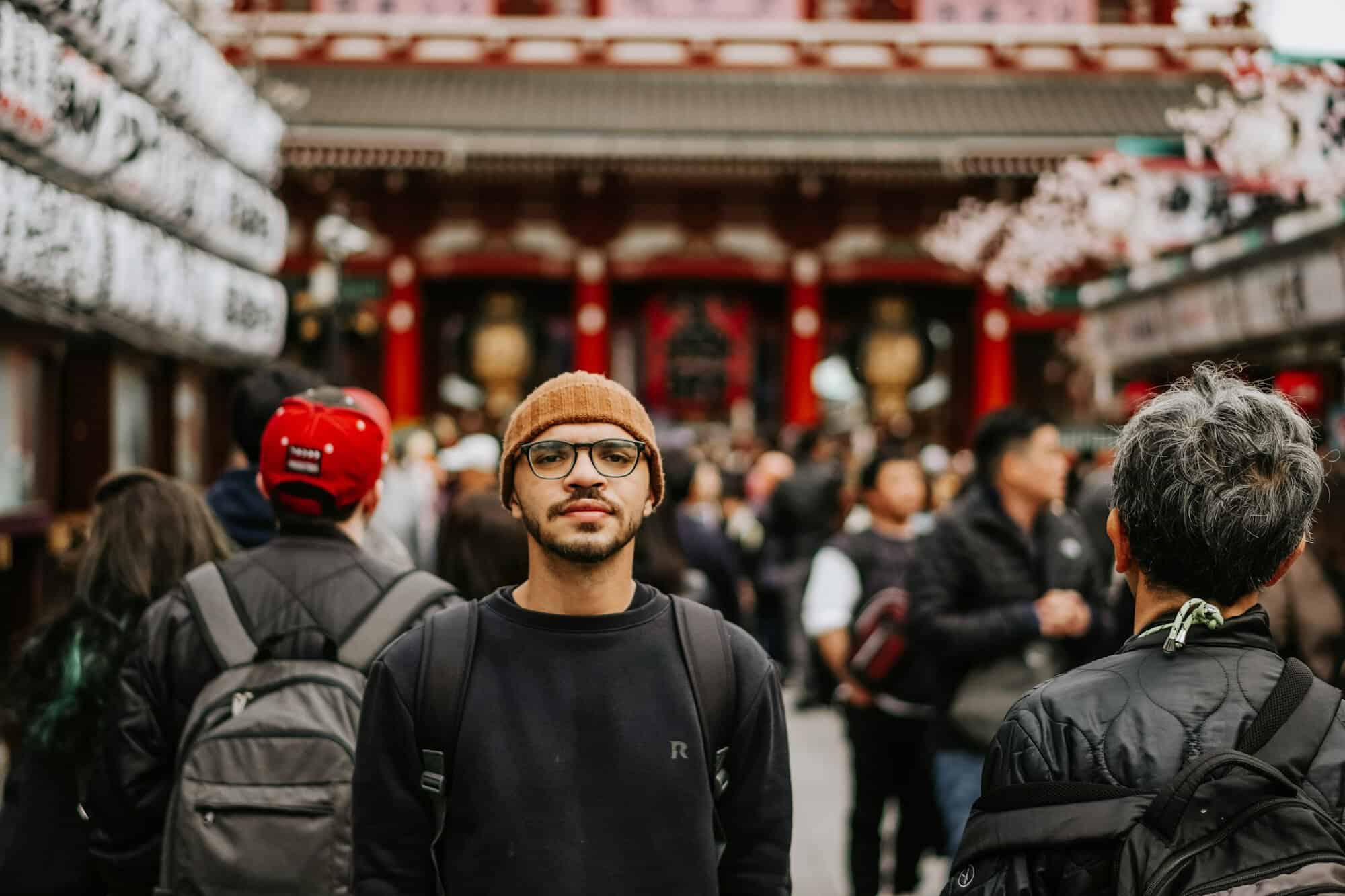 A smiling young man in glasses and a beanie standing in a lively Tokyo crowd