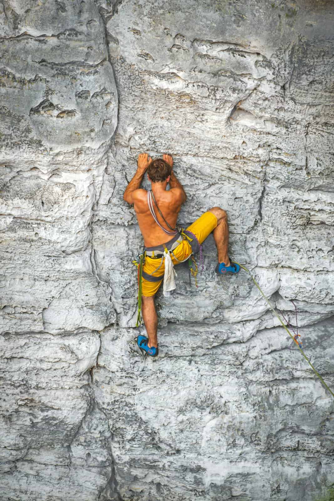A rock climber in yellow shorts reaching for the next hold on a pale stone wall, feeling stuck and moving one hold at a time