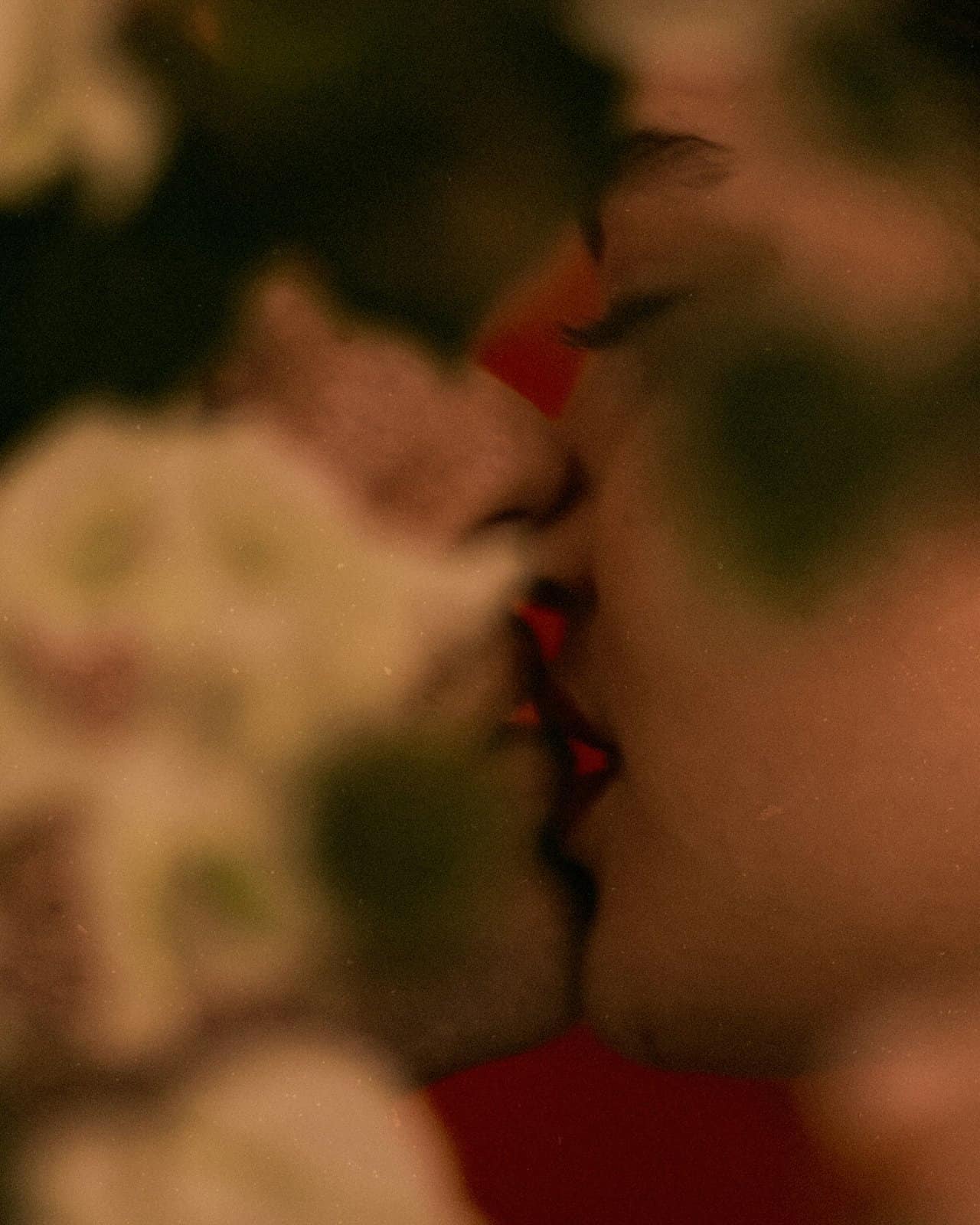 Two people sharing a soft kiss, out of focus behind white flowers in warm dark light, meaning in everyday closeness