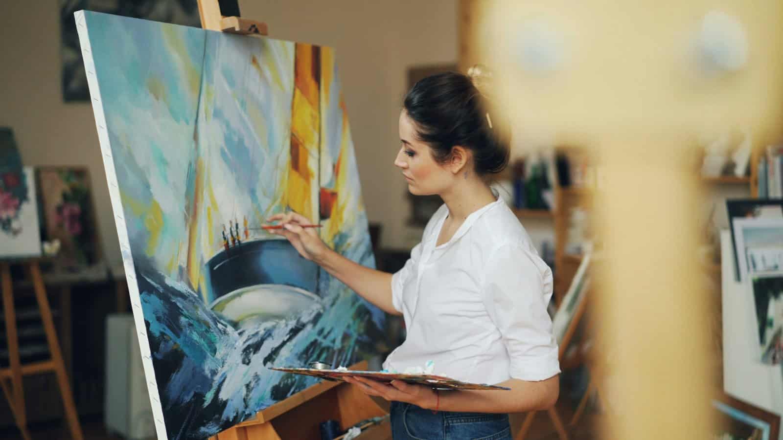 A woman in a white shirt painting a seascape at a large easel in a bright studio, fully absorbed in the present moment
