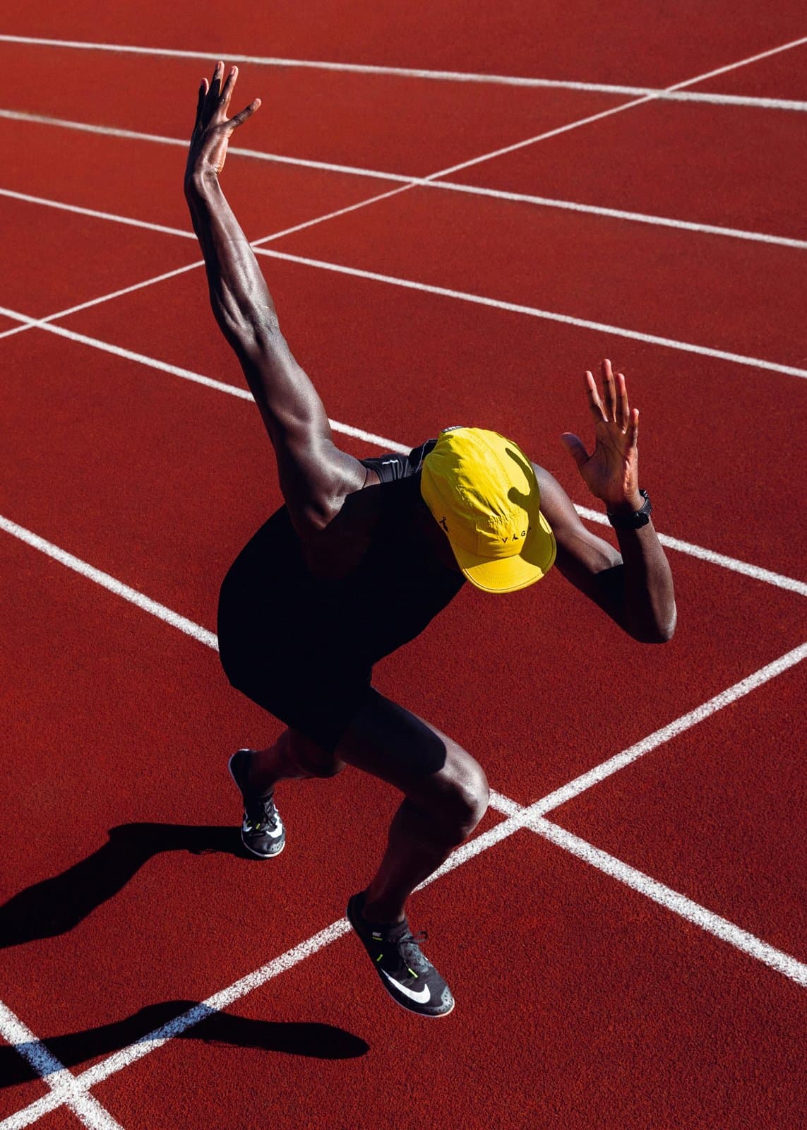 A sprinter in a yellow cap and black kit launching off the line on a red track, one arm raised, the first step of a fresh start