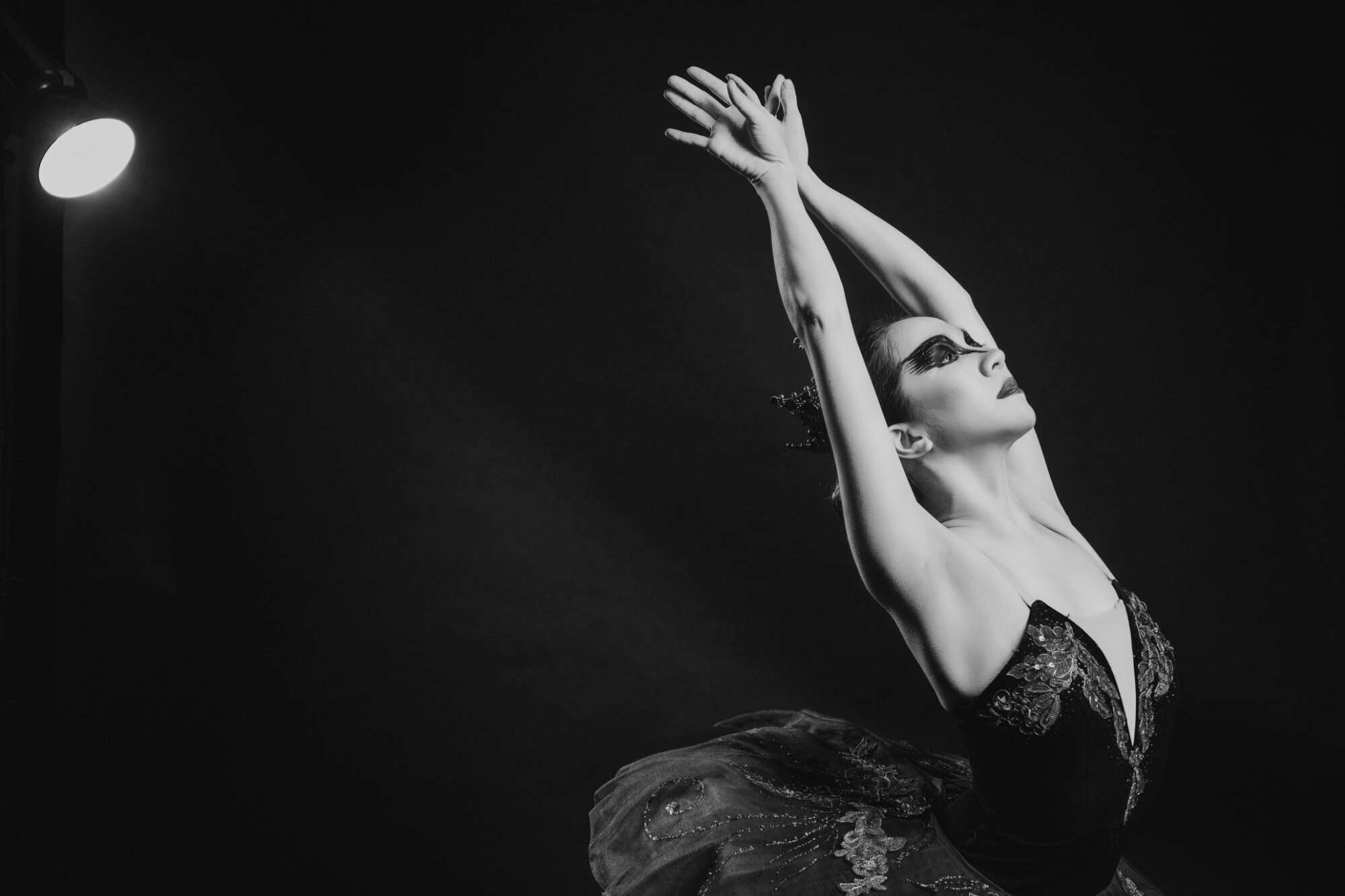 A ballerina in a black swan costume with arms raised overhead against a dark studio background, black and white, releasing the old role