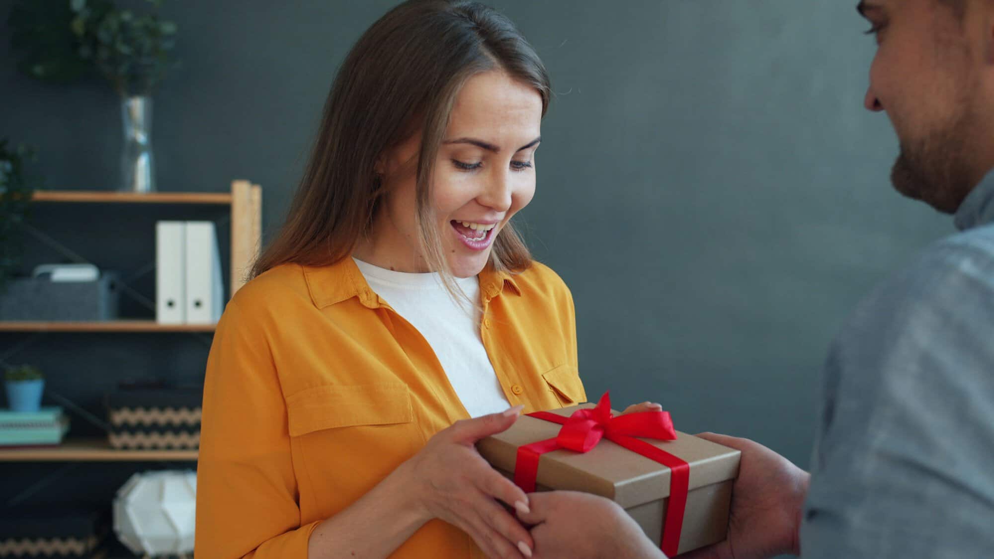 A smiling woman in a mustard shirt receiving a gift box tied with red ribbon