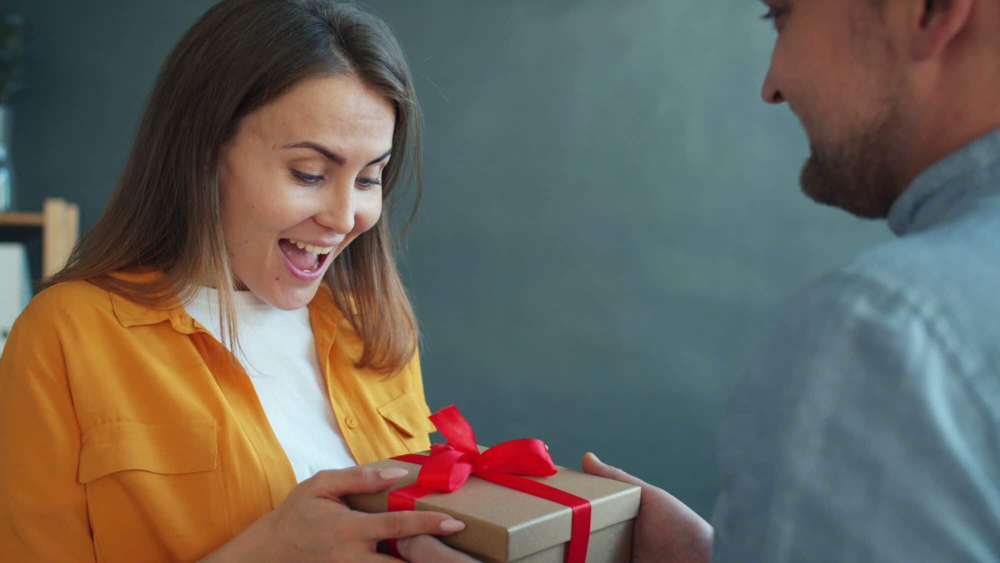 A delighted woman in a yellow shirt receives a wrapped gift with a red ribbon, a moment that Japanese gifts for mom are chosen for