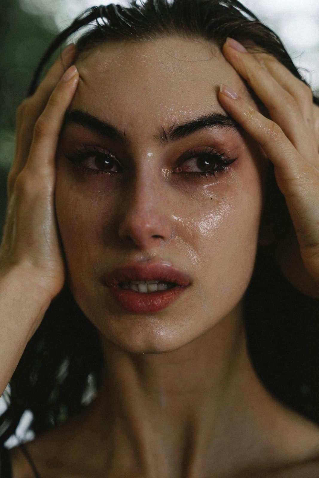 A young woman with wet lashes and both hands pressed to her head, overwhelmed by missing someone