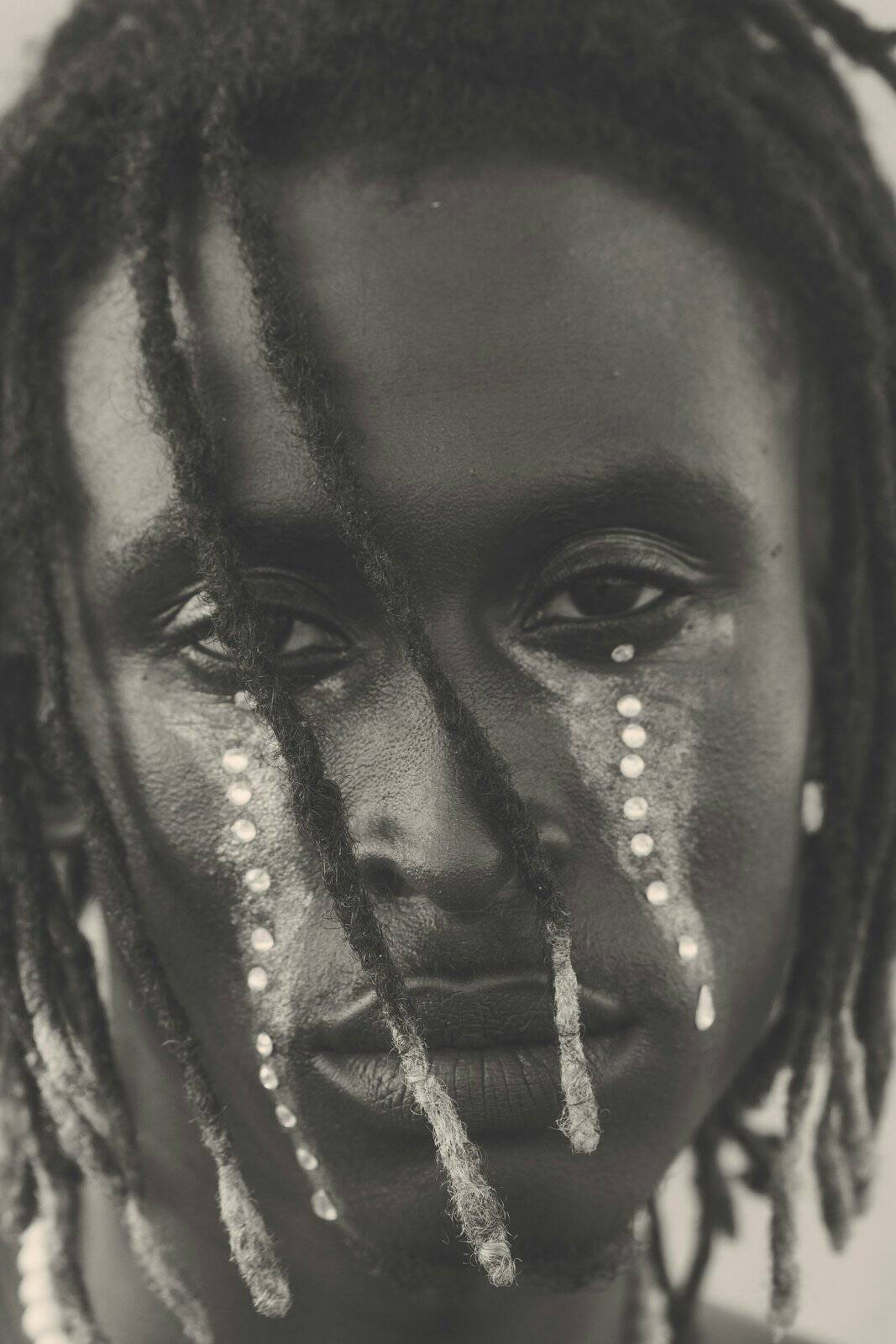 Black-and-white portrait of a young person with dreadlocks and pearl-like tears painted down both cheeks, calm eyes meeting the camera