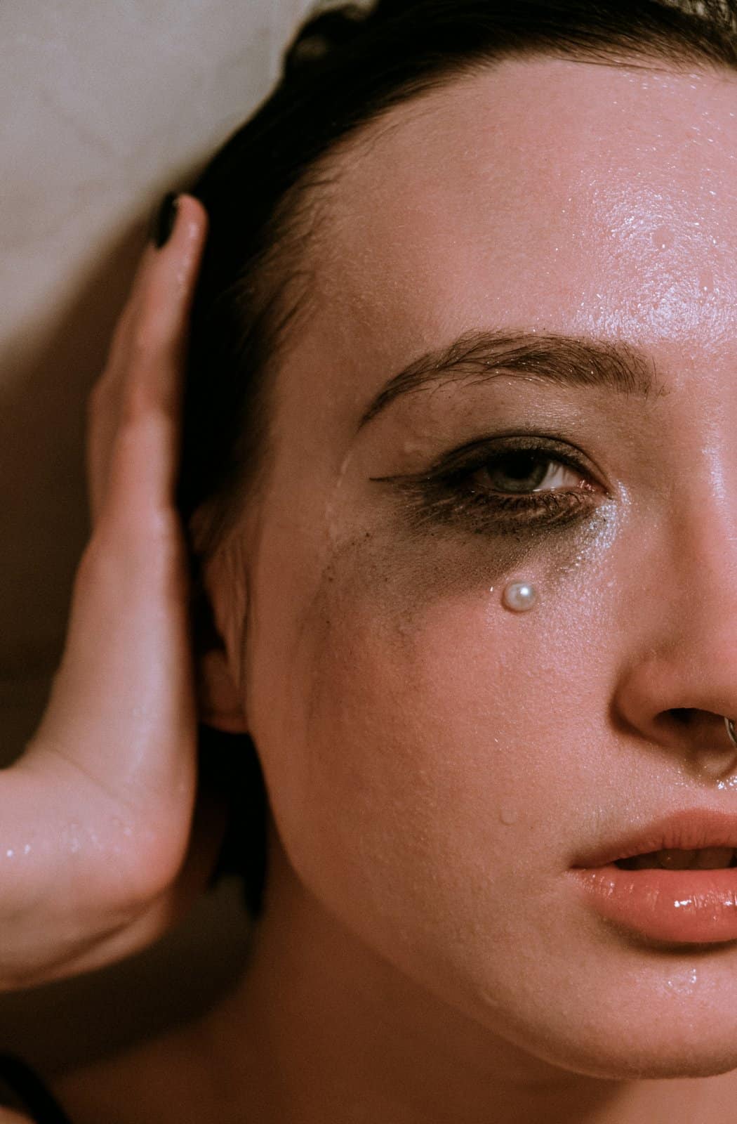 A young woman with smudged eyeliner and a single pearl bead resting on her cheek like a tear, one hand pressed to her ear