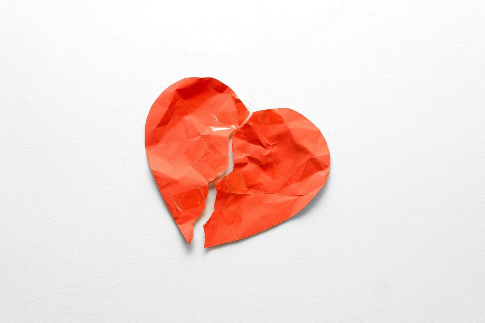 A crumpled red paper heart torn in two and held together with clear tape on a white wall, heartbreak at its plainest
