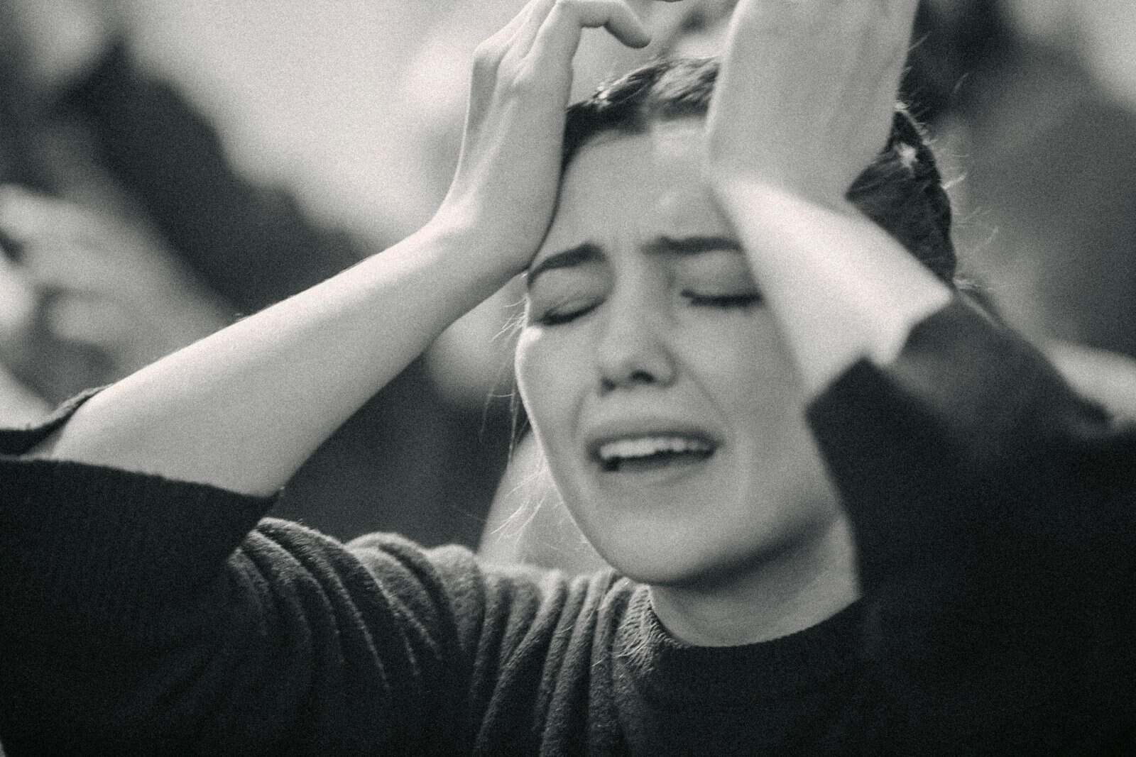 A woman in a dark knit sweater weeping with her hands raised to her head, photographed in black and white