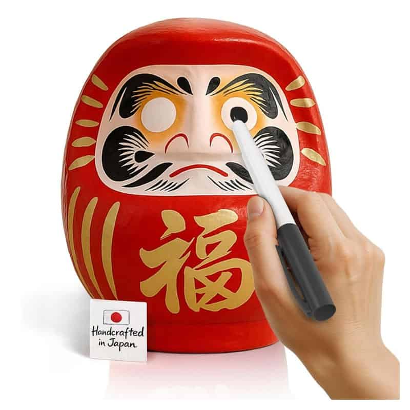 A red Takasaki daruma doll with blank eyes and a hand filling in one eye with a marker