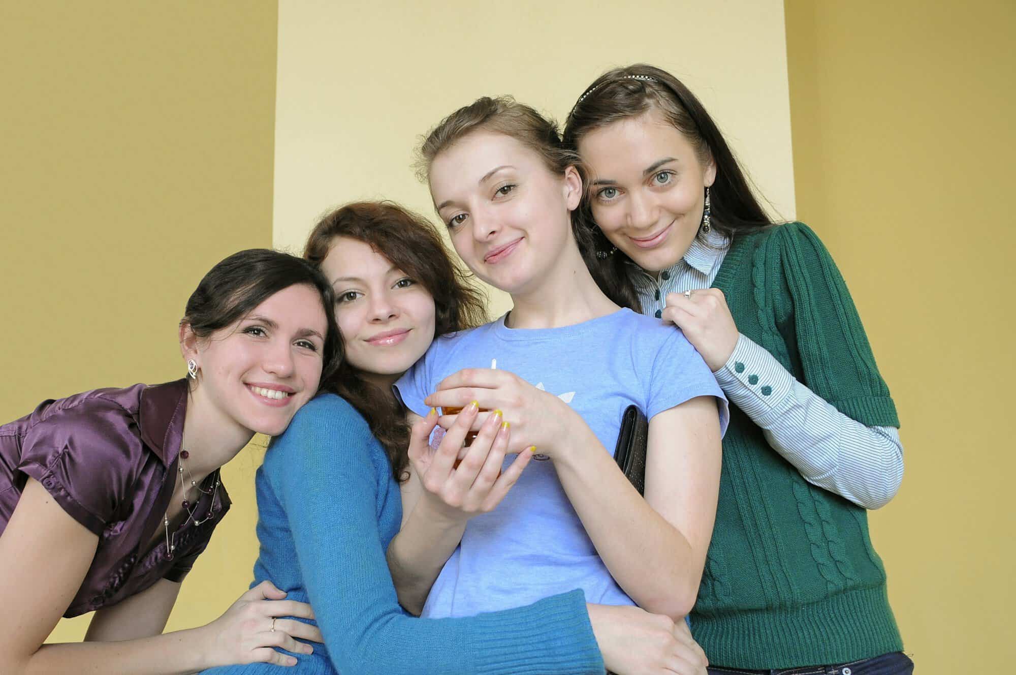 Four smiling young women hugging close together against a yellow wall